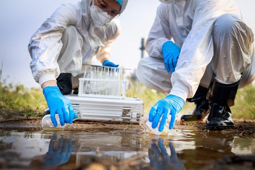 An operator in protective coveralls and gloves collecting a representative sludge sample into a bottle at the edge of a wastewater lagoon.
