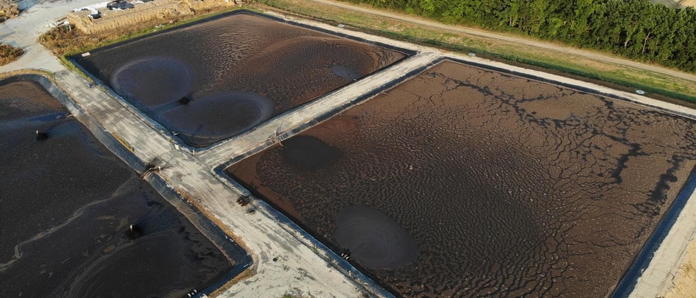 Aerial view of drawn-down lagoon cells with exposed dark sludge, divided by paved access roads — the state a mechanical clean-out works from.