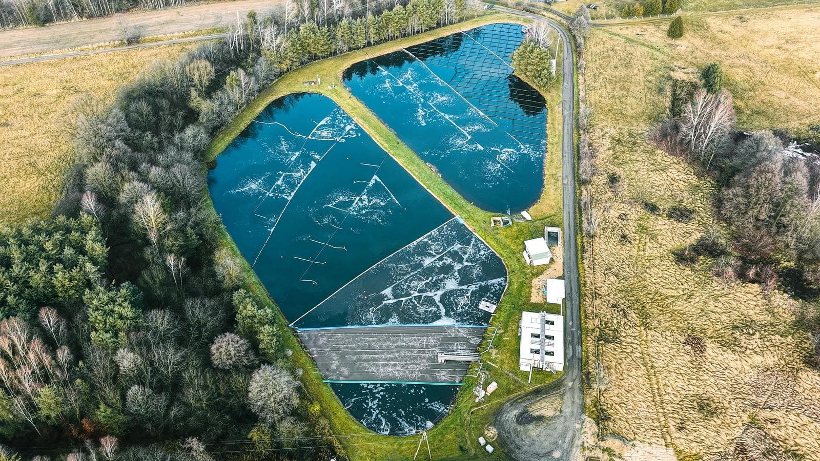 Aerial view of a multi-cell aerated wastewater treatment lagoon set in open countryside, with surface aeration visible across the water and grass berms between the cells.